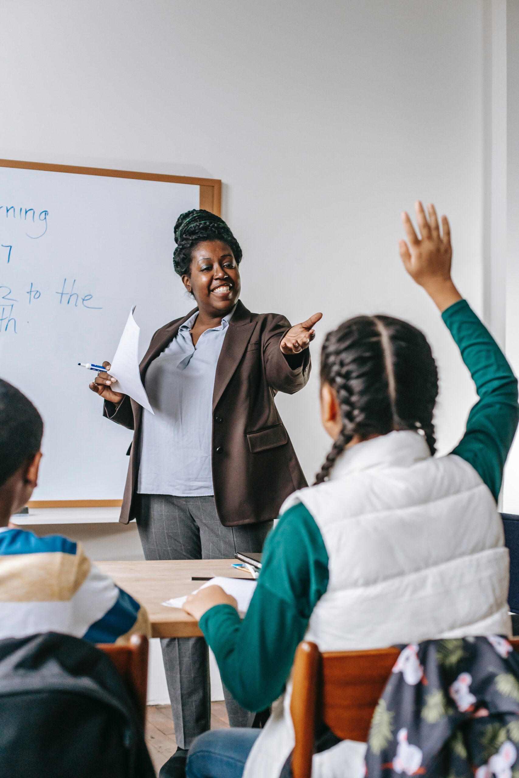 Professora negra, em frente a um quadro branco, sorrindo para aluna de costas com a mão levantada. A aluna está com 2 tranças, blusa verde e colete branco